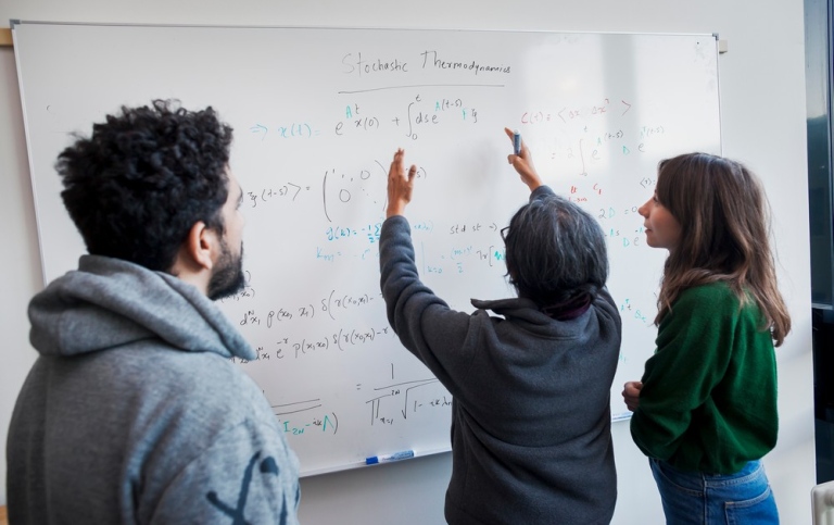 A physics teacher and two students in front of a whiteboard with stochastic thermodynamic equations