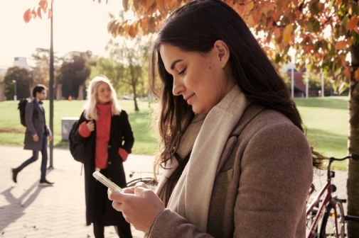 Student står utomhus och läser på mobiltelefon.