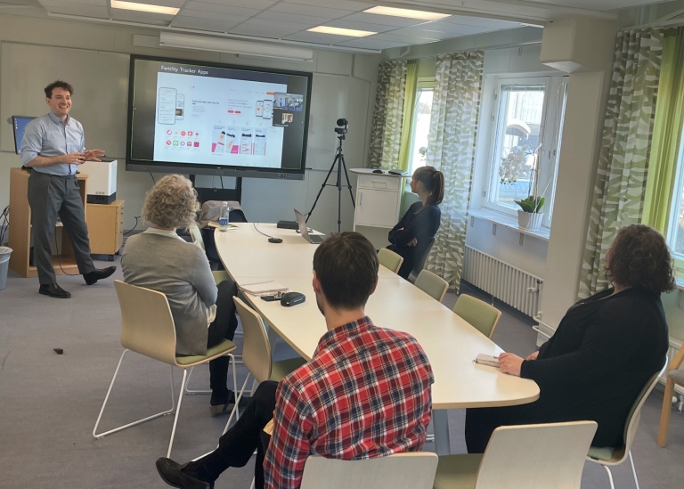 En forskare presentera sitt arbete framför en skärm, vissa åhörare finns på plats, andra online.