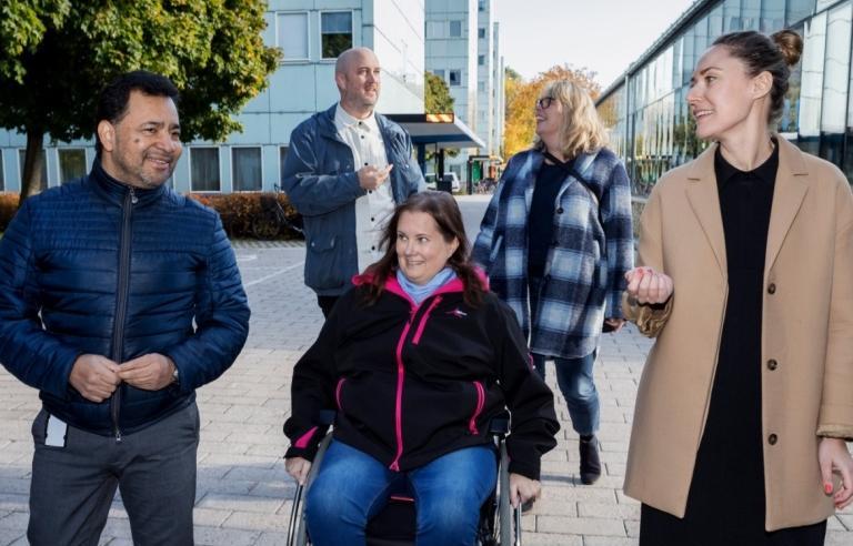 Två män och tre kvinnor pratar på campus. Kvinnan i mitten sitter i rullstol.
