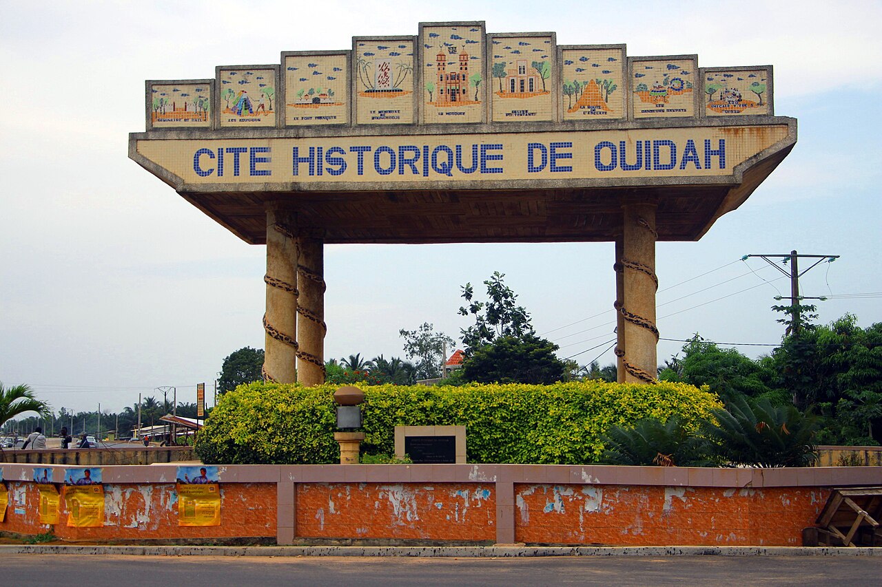 Entrance gateway to the historic city of Ouidah, with the words “CITE HISTORIQUE DE OUIDAH” displayed across the top. Green vegetation surrounds the gateway, with a road and power lines visible in the background.