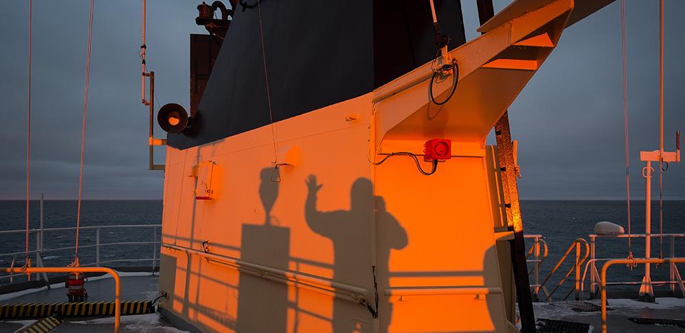 A man on the deck of an icebreaker ship at sunset waves at his own shadow