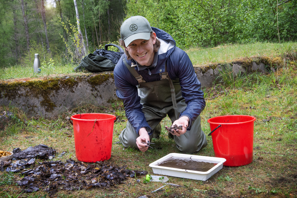 Man kneeling on the ground between two red buckets, holding a soil sample during fieldwork.