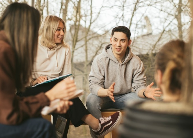 En grupp studenter sitter ute på campus. Fokus på kille som berättar något för de andra