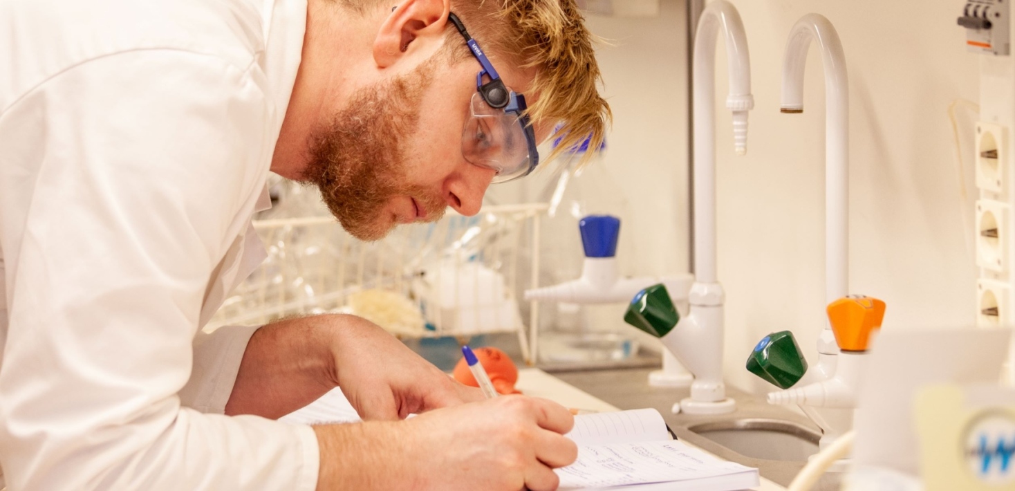 Person iklädd skyddsglasögon och labbrock som för anteckningar på lab.