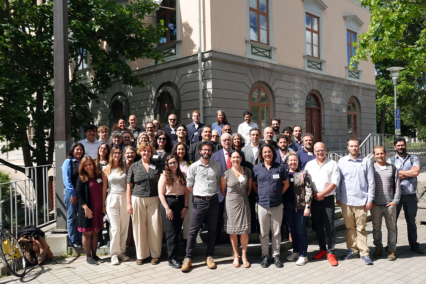 Group photo of numerous participants at the NOLAN 2026 conference organized by NILAS (Nordic Institute of Latin American Studies) at Stockholm University. The participants are gathered in several rows on outdoor steps in front of a light pink and grey building with large windows and arched doorways. The photo was taken on a sunny summer day with green trees in the background.