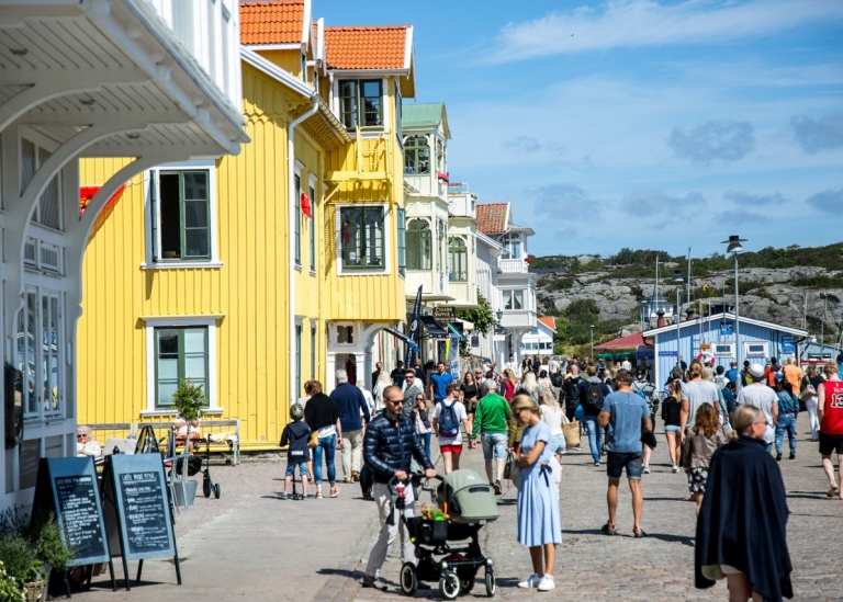 En solig sommardag i en svensk kuststad med färgglada trähus och människor som promenerar.