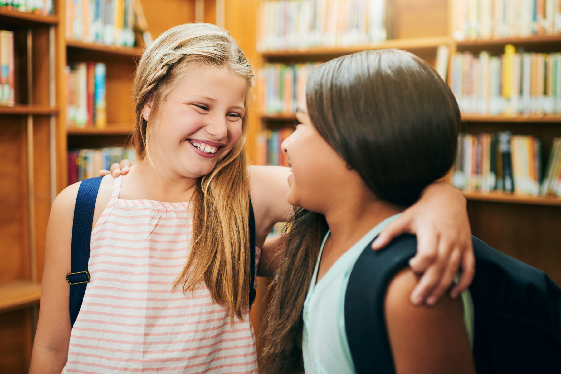 Two young schoolgirls standing with their arms around each other inside a school library during the day