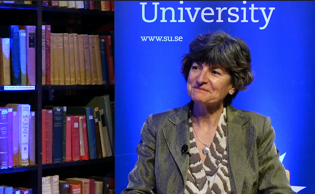 María Ángeles sitter framför en bokhylla fylld med böcker, bredvid en blå panel där texten ”University” och ”www.su.se” syns. Hon bär en grå kavaj och har en mygga fäst vid klädseln.