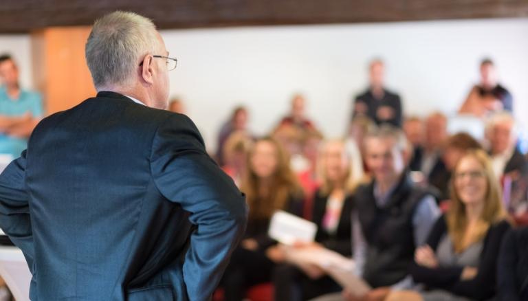 Äldre man står inför en publik och talar