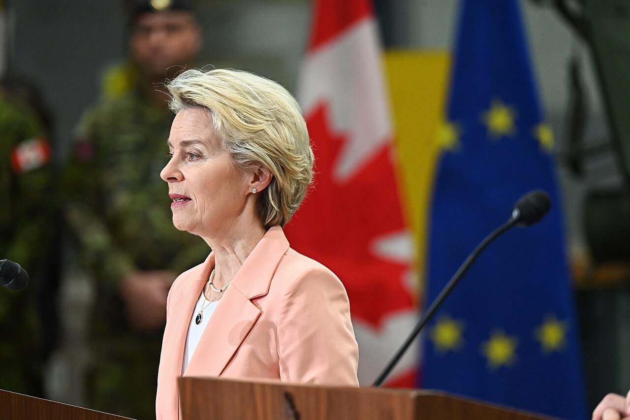 Ursula von der Leyen, ordförande för Europeiska kommissionen, talar vid ett podium under sitt besök i Kanada. Hon bär en ljus persikofärgad kavaj över en vit blus, med ett pärlhalsband. Bakom henne syns den kanadensiska flaggan (röd och vit med ett lönnlöv) och Europeiska unionens flagga (blå med gula stjärnor). En uniformsklädd medlem av de kanadensiska styrkorna står i bakgrunden.