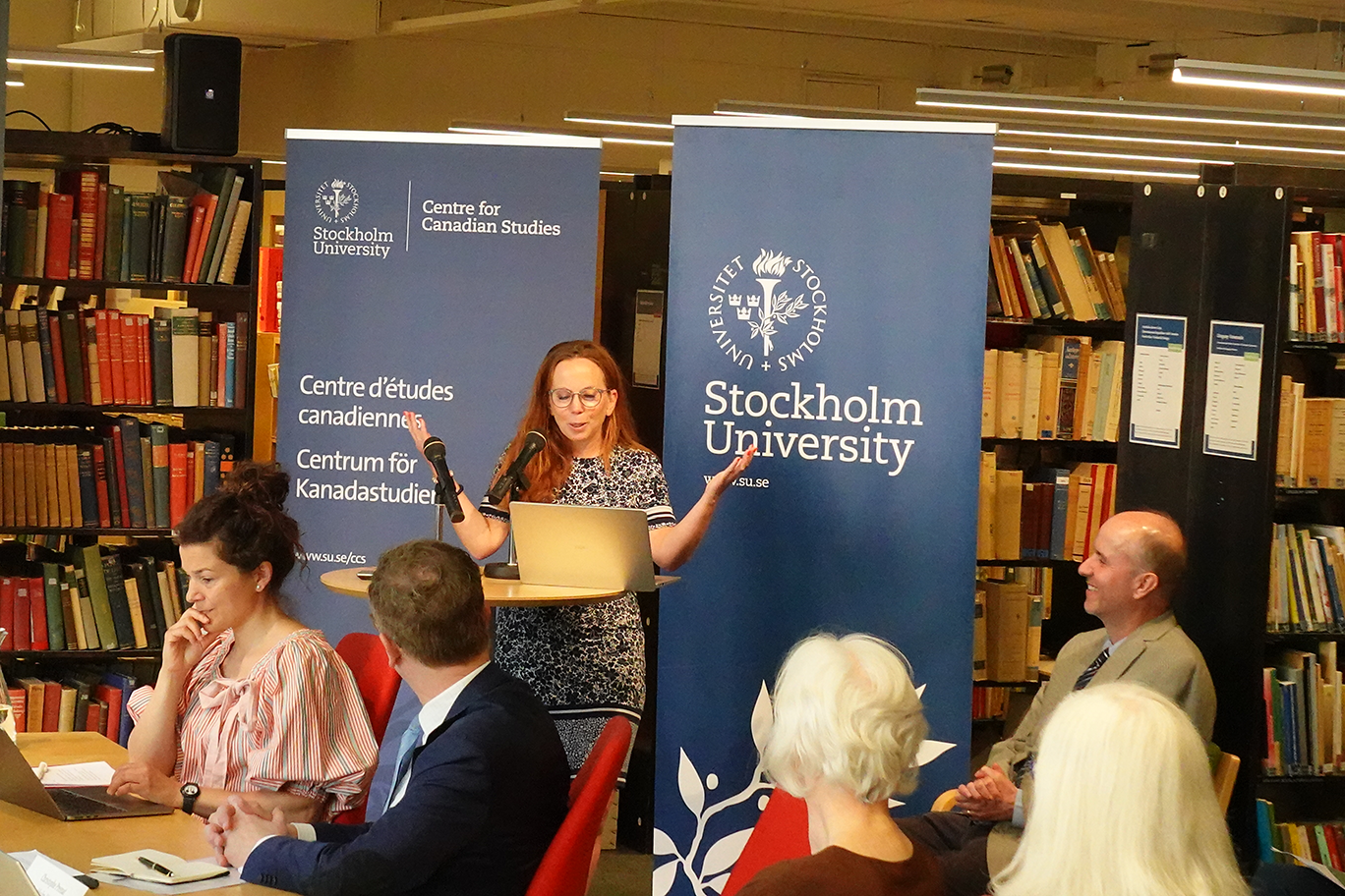 Conference participants at the Centre for Canadian Studies, Stockholm University. A woman speaks at a podium in front of banners bearing the university's and the centre's logos, surrounded by attendees in a library setting.