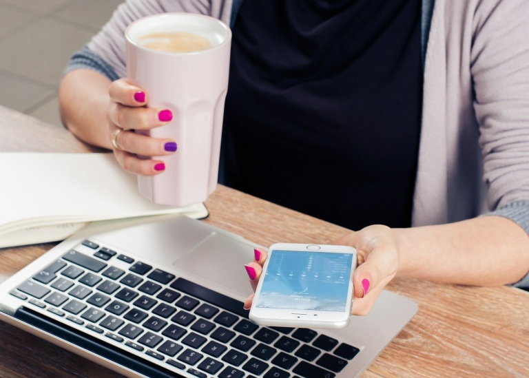 Person vid skrivbord med dator, mobil, anteckningsbok och lattemugg. Foto: Firmbee från Pixabay.