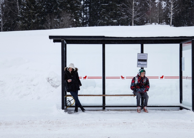 Två personer väntar vid en snötäckt busshållplats, en använder mobilen, den andra har hörlurar.