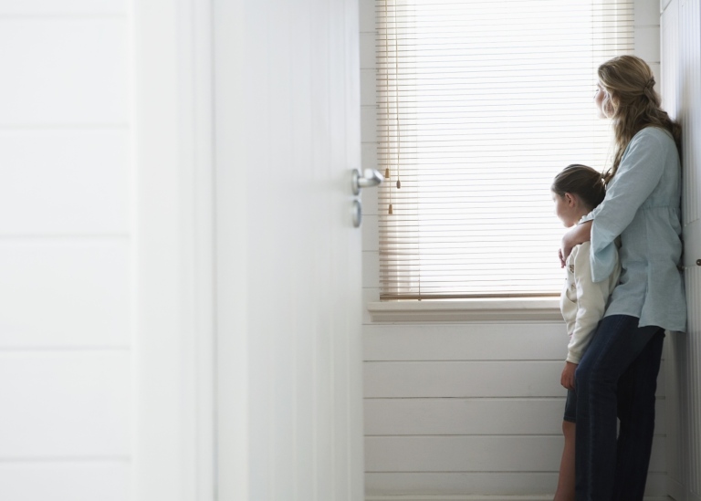 Sidovy av mor och dotter som tittar ut genom ett fönster med persienner