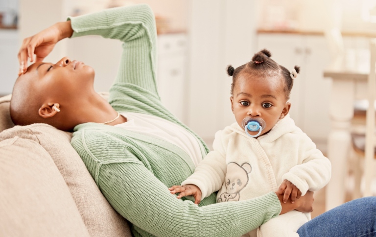 A tired mother reclines on a couch with her hand on her forehead while holding her baby-