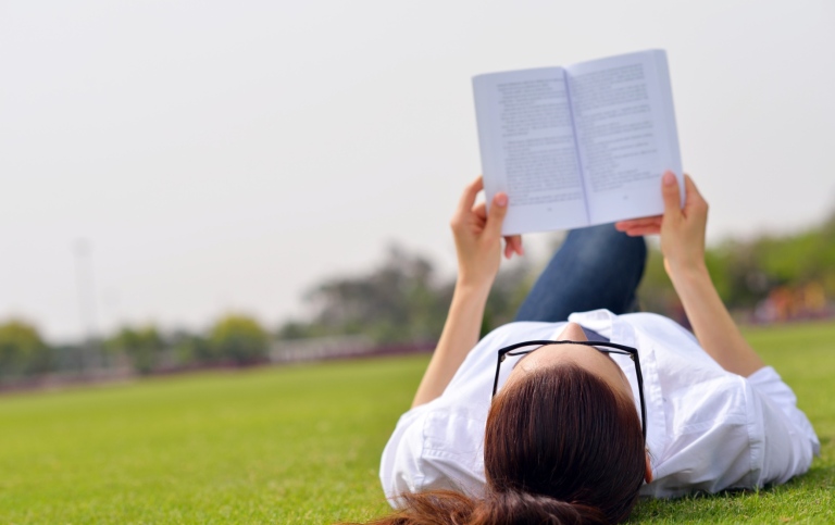 Young woman lying on her back in a park reading a book.