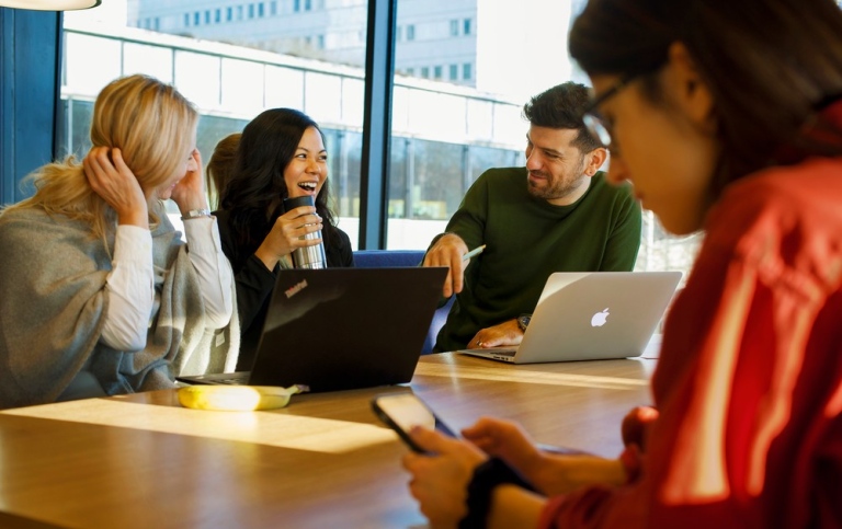 Tre studenter sitter vid laptops och pratar och skrattar. I förgrunden en student med mobiltelefon.