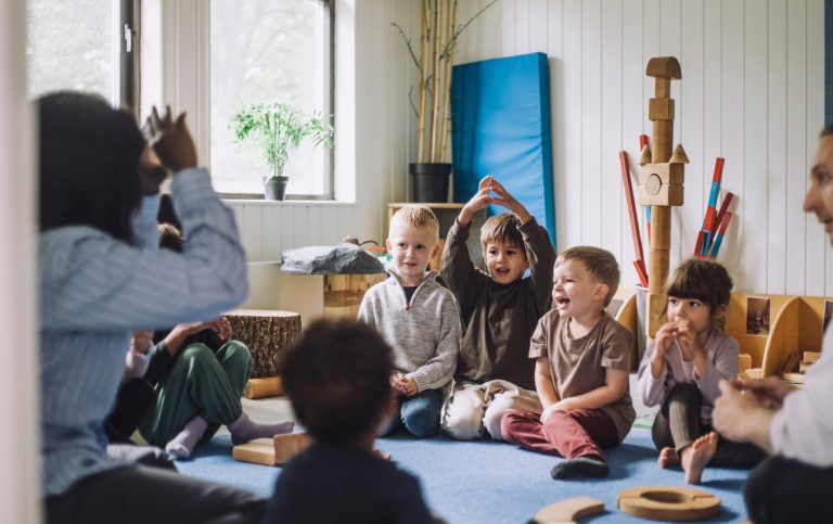 Två förskolärare och en barngrup har sångsamling inomhus.