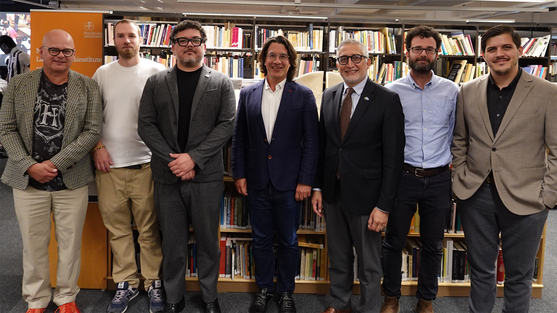 Seven people stand in front of bookshelves in a library or reading room. In the background, an orange sign displays the text “Stockholm University” and “Nordic Latin American Institute”.