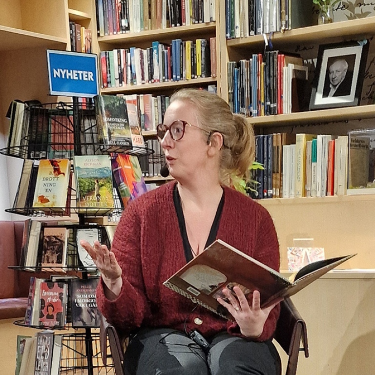 Sara Van Meerbergen pratar och håller i en stor bilderb. I bakgrunden bokställ och bokhyllor.