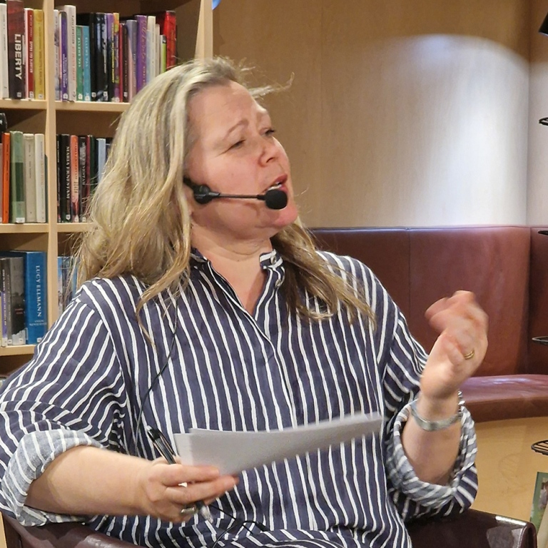 Elina Druker tittar på övriga i panelen, pratar i headset, håller i papper och gestikulerar.