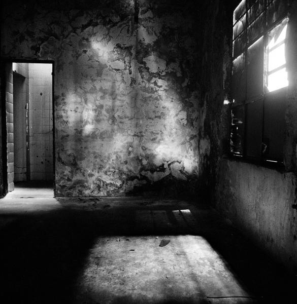 Black and white photograph of a dilapidated room with peeling walls, sunlight streaming through a barred window, casting shadows on the worn floor. A doorway leads to a tiled corridor.