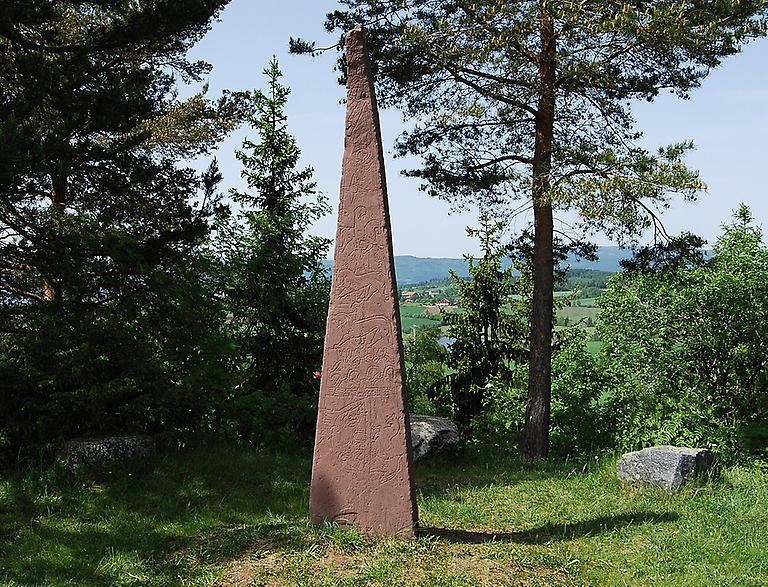Hög smal konformat röd runsten på gräsmatta omgiven av träd och vanliga stenar. Landskap i bakgrunden.