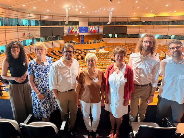 A group of seven people standing in a row inside the European Parliament hemicycle in Brussels, with rows of orange seats and the EU flag visible in the background. The group includes four women and three men dressed in business-casual attire, most wearing name tags, posing for a photo on the upper level overlooking the debating chamber.