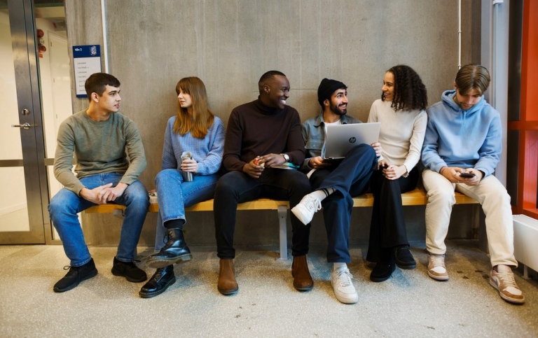 En grupp studenter sitter på en bänk inomhus och pratar.