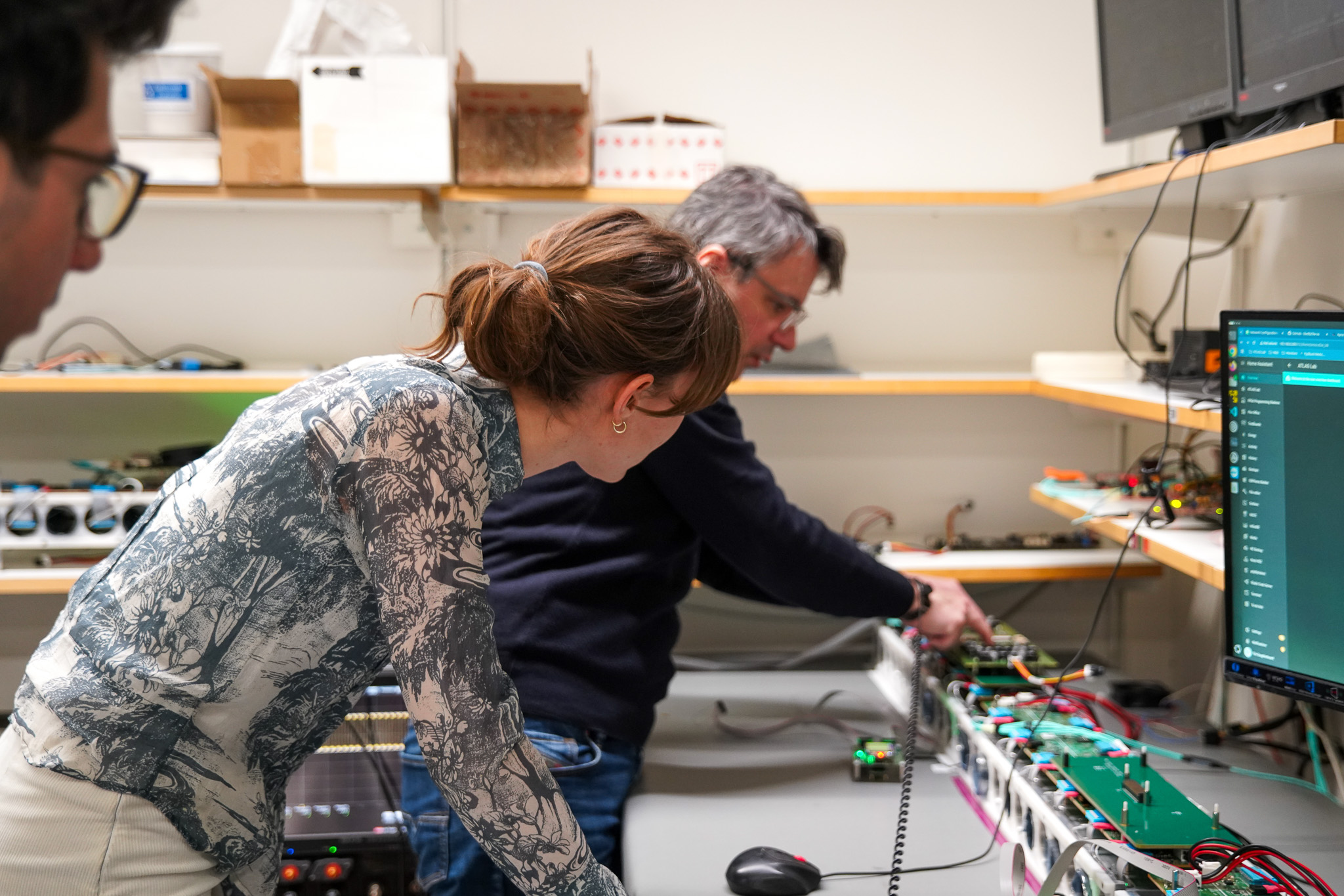 PhD students Federico Bonaldo and Julia Salwén work at a lab test bench alongside Professor Christophe Clément, with circuit boards visible.
