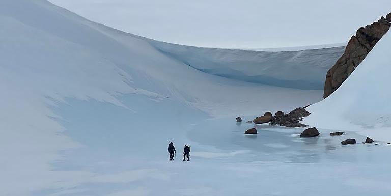 Two persons are walking on a snow field. Windtunnels form along the flanks of mountain summits that tower above the East Antarctic Ice Sheet.