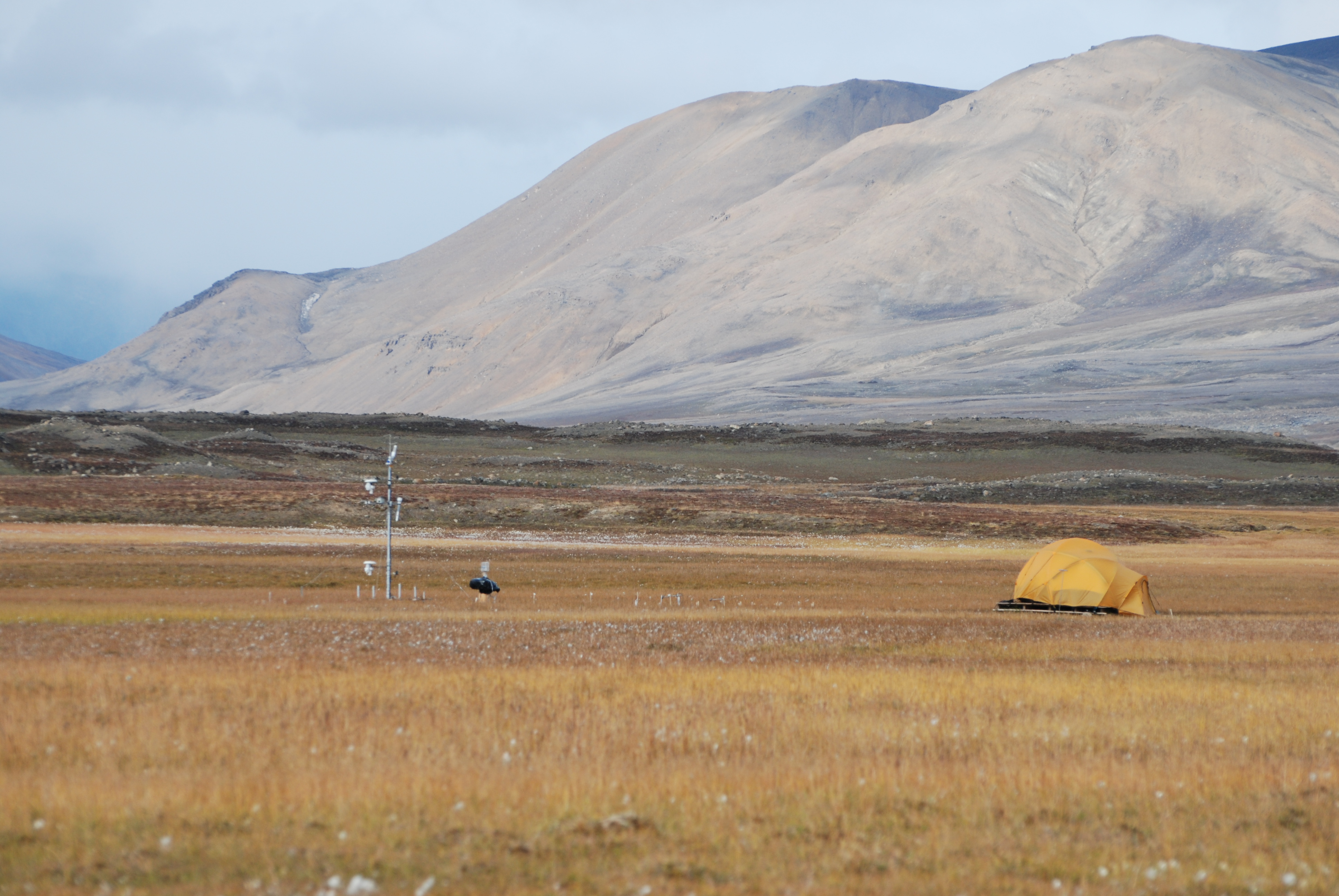 Mountains without trees in the background and a field of dry grass in the foreground. A yellow tent stands on the right, and a methane measurement tower is on the left.