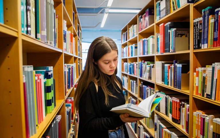 En person står och bläddrar i en bok i ett bibliotek.