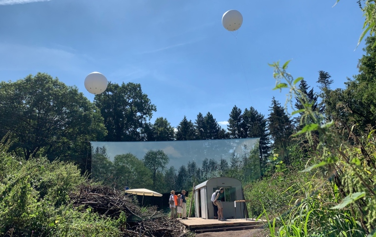 Framför kanten till en skog hänger en duk med en bild av samma skog. Konstverk.