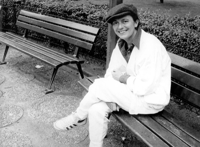 Bilden visar Monique Wittig (1935–2003), den franska författaren, feministiska teoretikern och lesbiska aktivisten, i ett svartvitt porträtt fotograferat av Colette Geoffrey.I bilden sitter Wittig på en parkbänk i en utomhusmiljö, troligen i en park eller trädgård. Hon är klädd i en ljus jacka, ljusa byxor och en mörk keps. På fötterna bär hon Adidas-träningsskor med de karakteristiska tre ränderna. Hon sitter avslappnat med benen i kors och ler mot kameran, vilket ger bilden ett informellt och personligt uttryck. Bakgrunden visar ytterligare bänkar, buskar och ett stenlagt underlag.Fotot fångar en vardaglig, nästan lekfull stämning – en kontrast till hennes intellektuella och revolutionära persona som en av nyfeminismens centrala teoretiker i Frankrike. Wittig är mest känd för sin banbrytande roman Les Guérillères (1969) och sina teorier om heteronormativitet och "könet som klass", som har haft enormt inflytande på feministisk och queer-teori.Bilden är licensierad under CC BY-SA 4.0 och tillgänglig via Wikimedia Commons, publicerad av Succession littéraire de Monique Wittig (Monique Wittigs litterära arv).