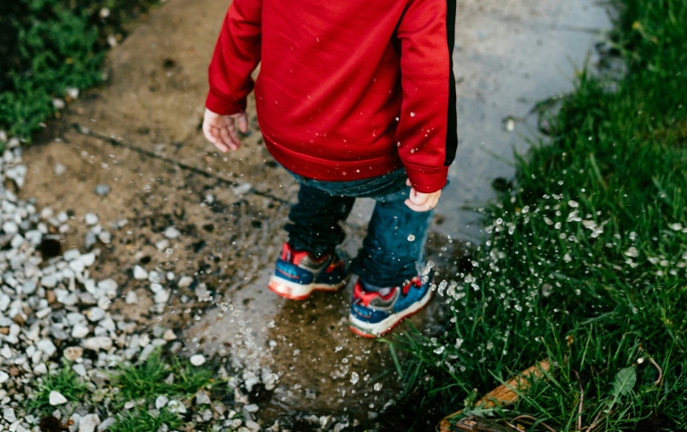 young child wearing a red jacket, blue jeans, and colorful sneakers jumps into a puddle.