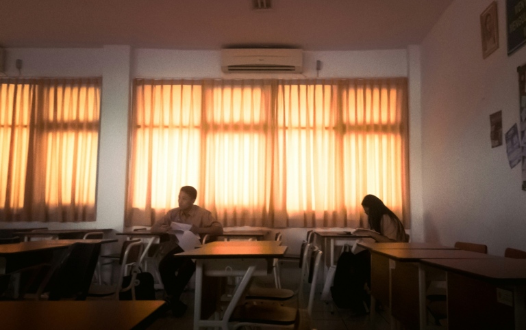 Two students sit apart in a dimly lit classroom.