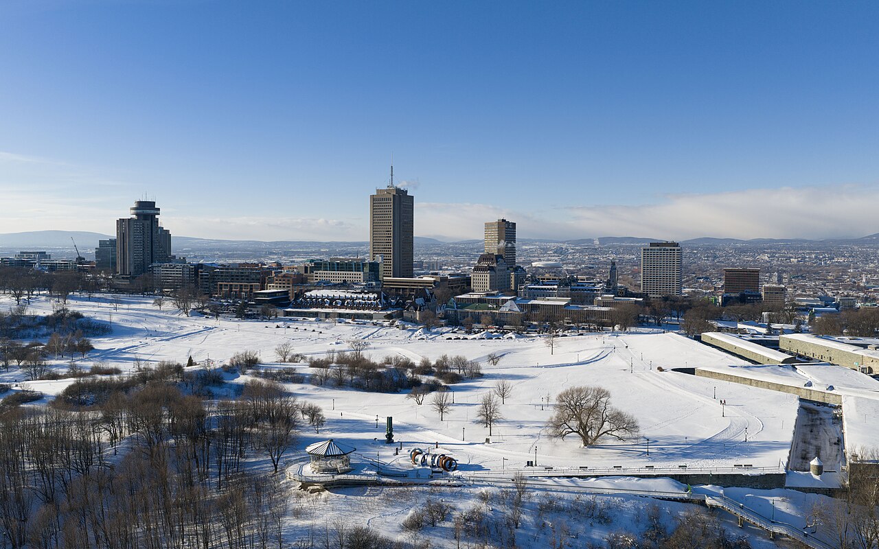 Panoramavy över Québec City under vintern, med snötäckt parkmark i förgrunden och stadens skyline med höghus i bakgrunden under klarblå himmel.The skyline of Quebec City, Canada, pictured in January 2026.