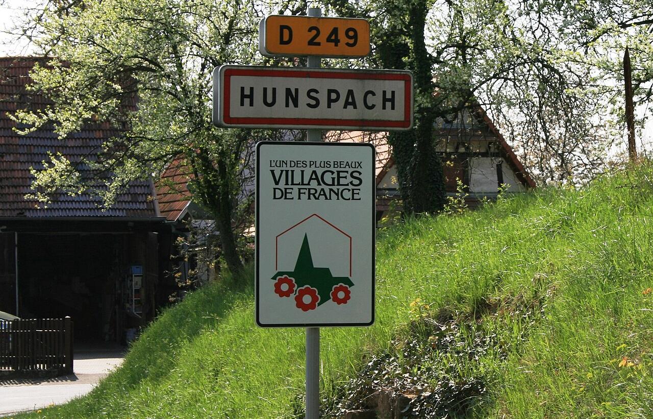 Vägskylt vid infarten till byn Hunspach i Frankrike, med vägnummer D 249, ortnamn och skylt för "Les Plus Beaux Villages de France". I bakgrunden syns blommande träd och traditionella hus med tegeltak. Bild: Lutz Fischer-Lamprecht, CC BY-SA 4.0 , via Wikimedia Commons