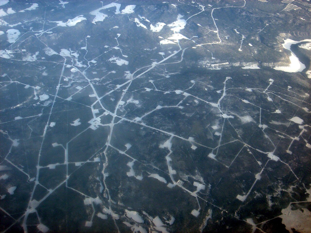 Flygfoto över ett oljefält i Alberta, Kanada, som visar ett nätverk av isiga vägar och borrningsplatser utspridda över ett snötäckt och skogklätt landskap.