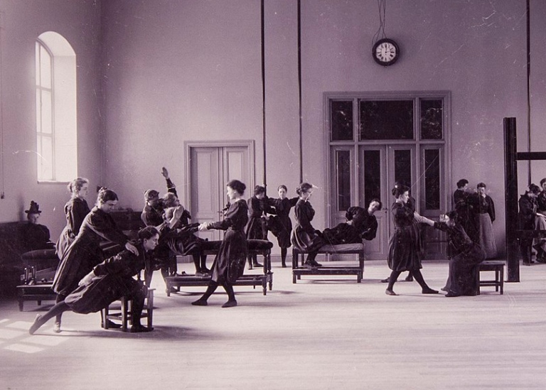 Svartvitt foto. Kvinnor i gymnastikhall gör gymnastiska övningar i gammaldags mörka kläder.