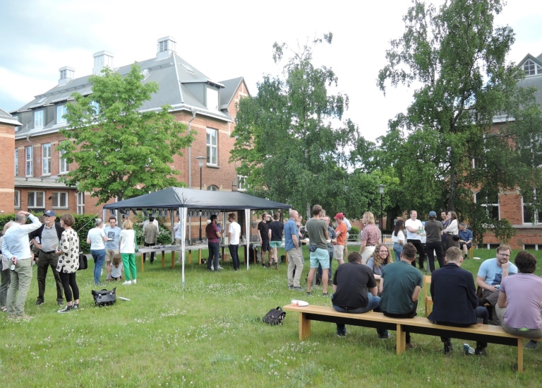 Sommarfest utomhus, folk som sitter på bänk och andra som står i olika grupper på gräsmatta.