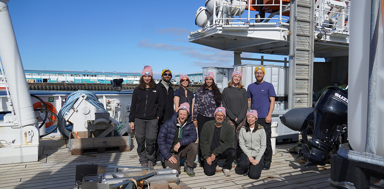 Researchers onboard a research vessel pose for a group photo, wearing expedition beanies in pink and yellow, each featuring the expedition logo.