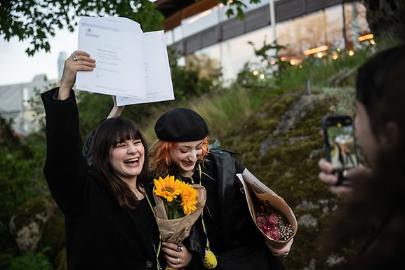 Two students with flowers smiling widely and holding up a diploma.