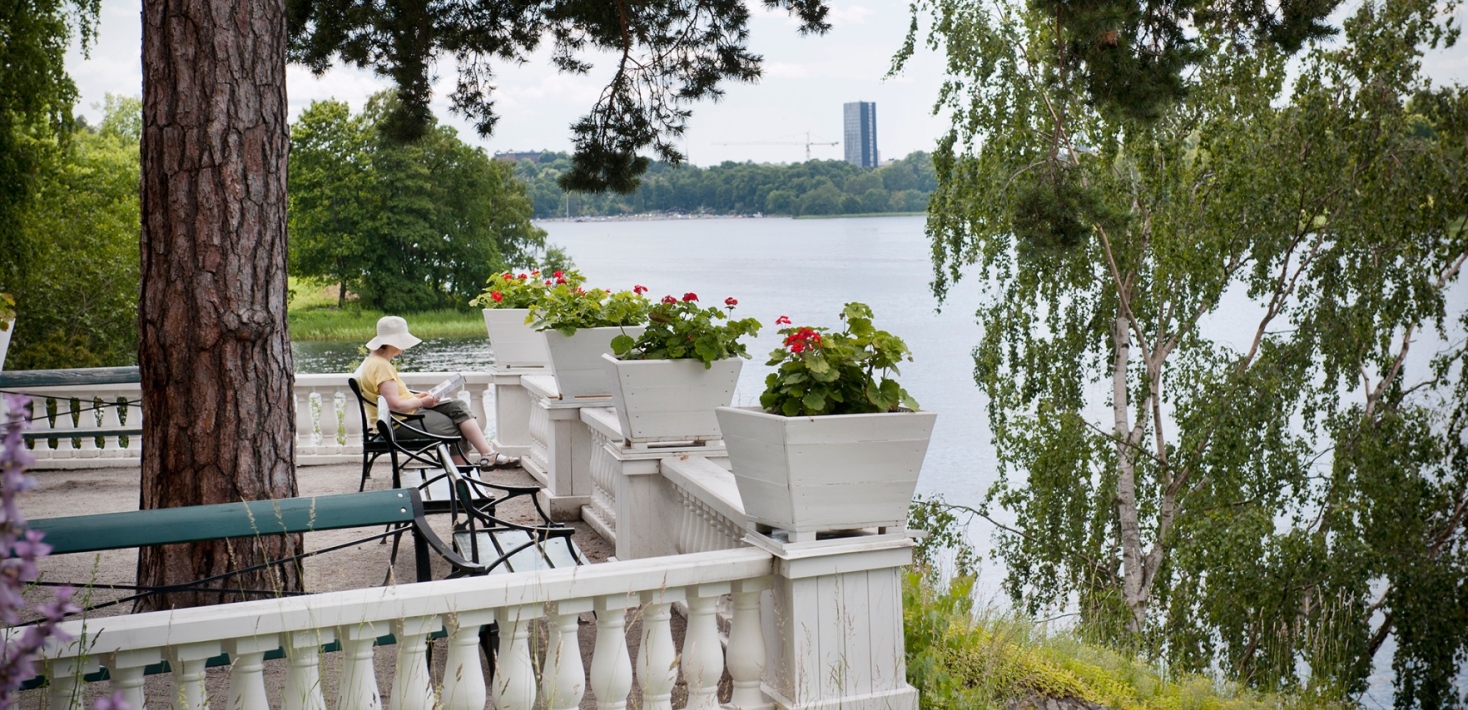 Italienska terrassen med vit balustrad. En kvinna sitter på en bänk och läser. Vy över Brunnsviken.