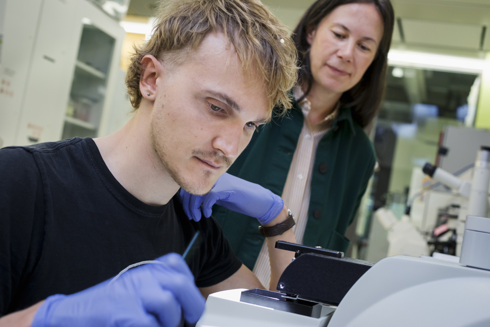 A woman stands behind a man in a lab. The man is wearing lab safety gloves and looks at a scientific instrument.