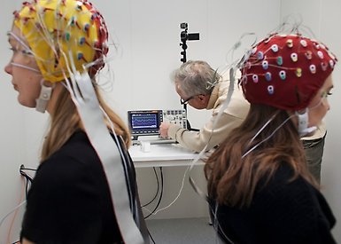 Två studenter med EEG-elektrodmössor på huvudet. I bakgrunden en forskare. Foto: Jens Olof Lasthein