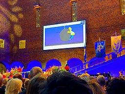 Bildskärm i Blå hallen, blommor, publikhav. På skärmen ser man Homer Simpson klamra sig fast vid en stor rivningskula i luften.
