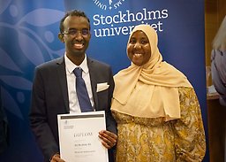 Manlig student i mörk kostym håller i diplom bredvid kvinna i gulmönstrad klänning och gul hijab